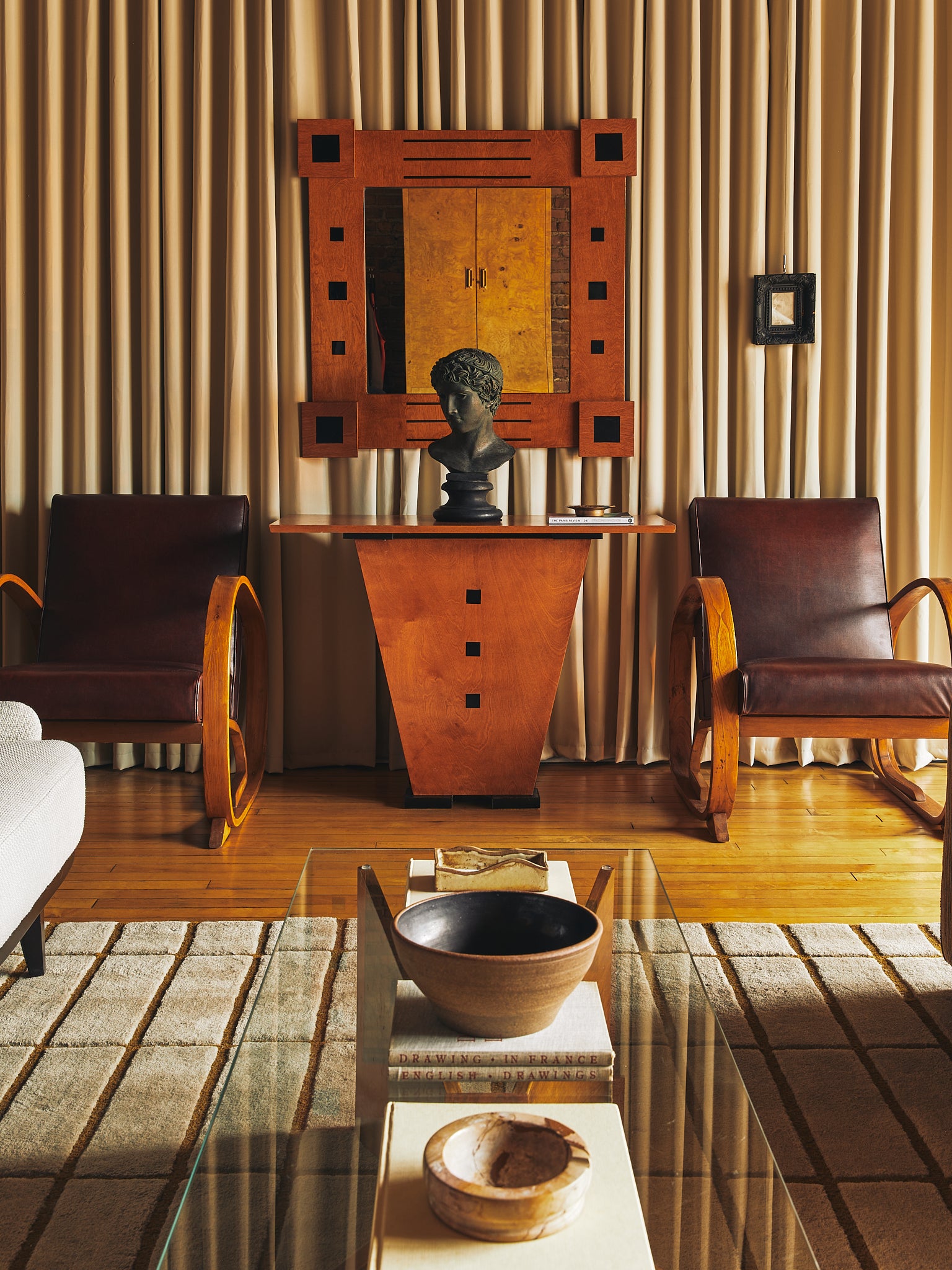 A mid-century modern living room featuring two brown leather rocking chairs with wooden frames, a matching wooden console table with a bust sculpture, and a large geometric mirror on a background of beige pleated curtains.