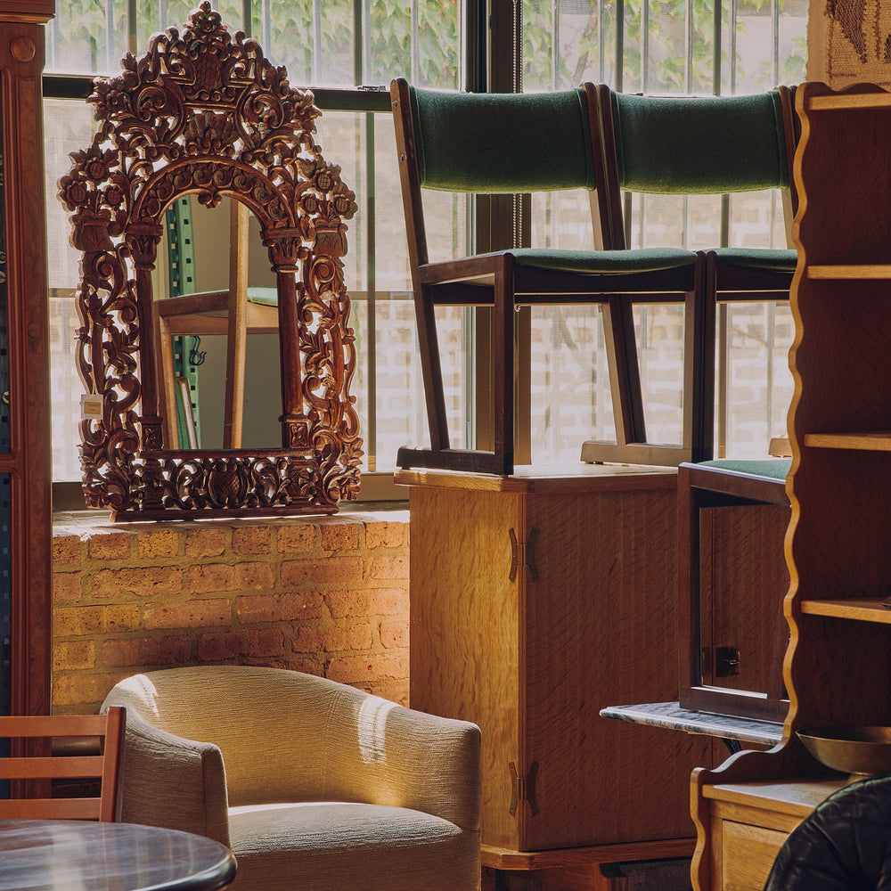 A styled interior scene featuring a carved dark wood mirror, a beige upholstered armchair, green cushioned chairs, wooden cabinets, and a dark leather chair in a sunlit room with brick walls and large windows.