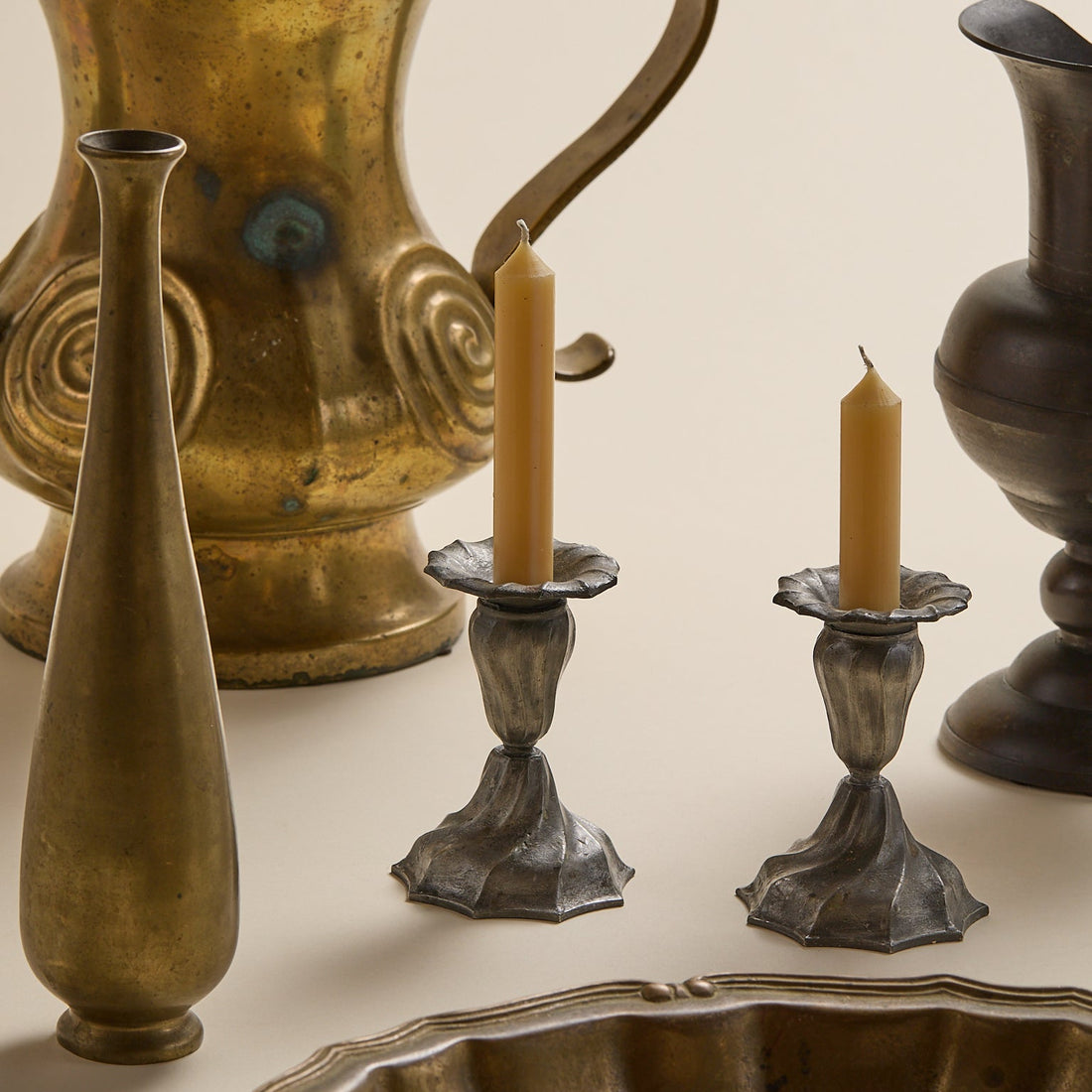 A styled collection of vintage brass and pewter home decor, including a tall slender vase, a candle holder with a taper candle, a matching pedestal candle holder, a large decorative bowl, and a textured metal pitcher, arranged on a warm cream background.