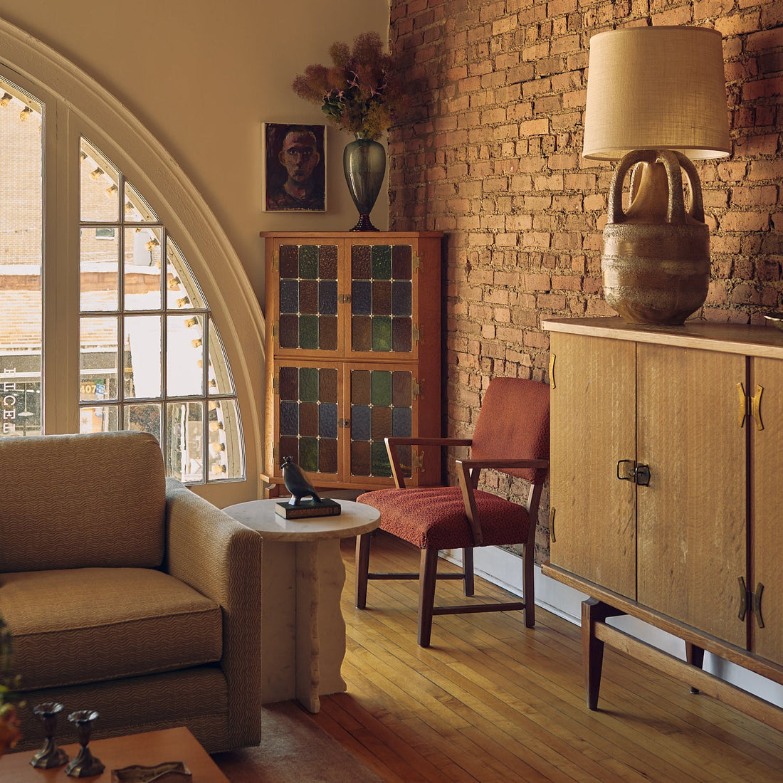 A warm, rustic living room corner featuring a brick wall, a mid-century wooden cabinet with a matching table lamp, a red upholstered accent chair, and a beige armchair.