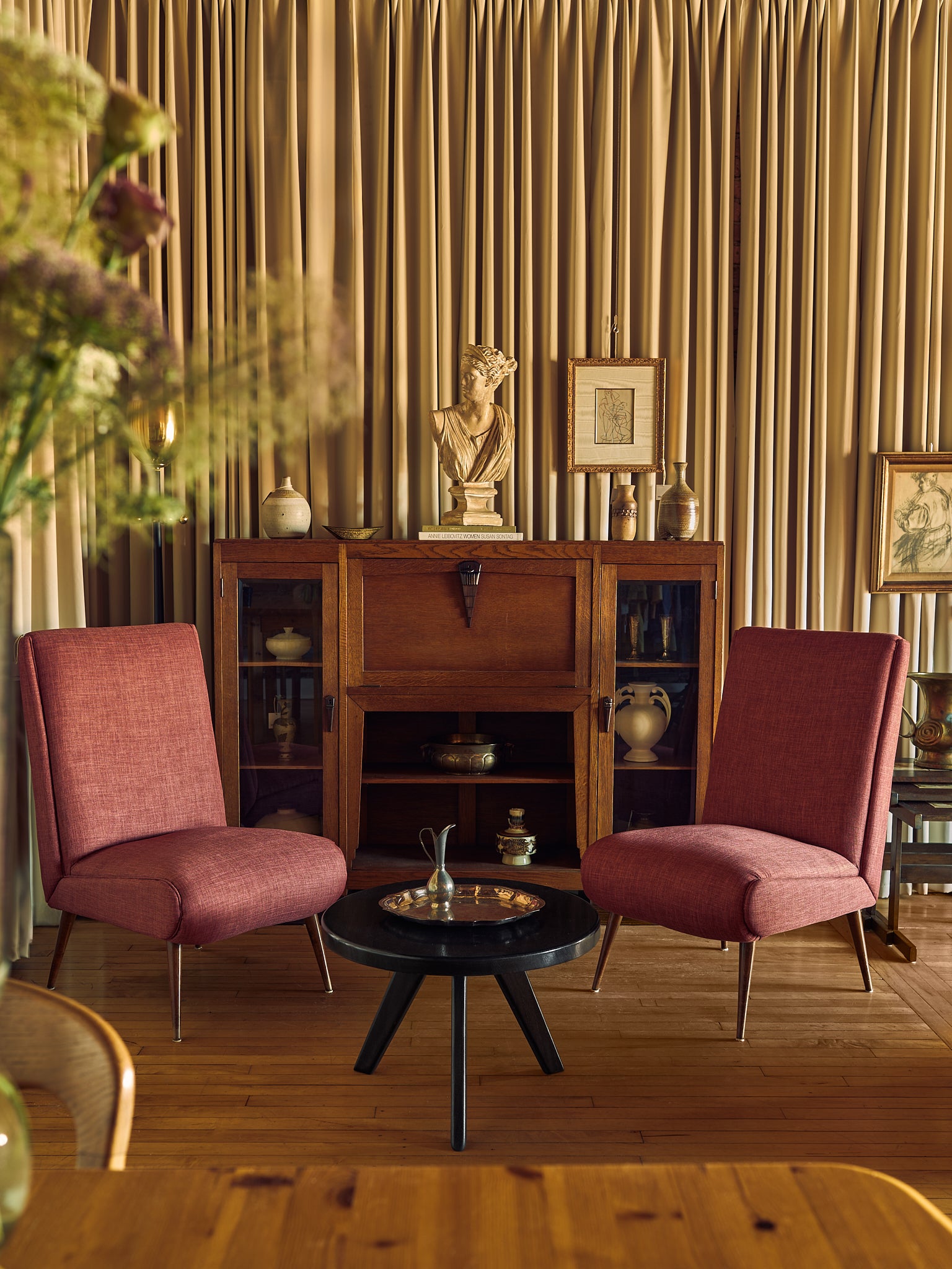 A styled interior scene with two pink upholstered armchairs facing a small round black coffee table, set in front of a wooden cabinet against a backdrop of pleated gold curtains.