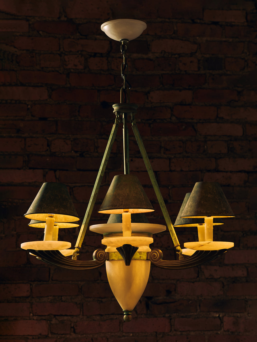 A vintage-style five-light chandelier with dark green metal arms, dark green fabric shades, and warm cream-colored glass shades, hanging against a dark red-brown brick wall.