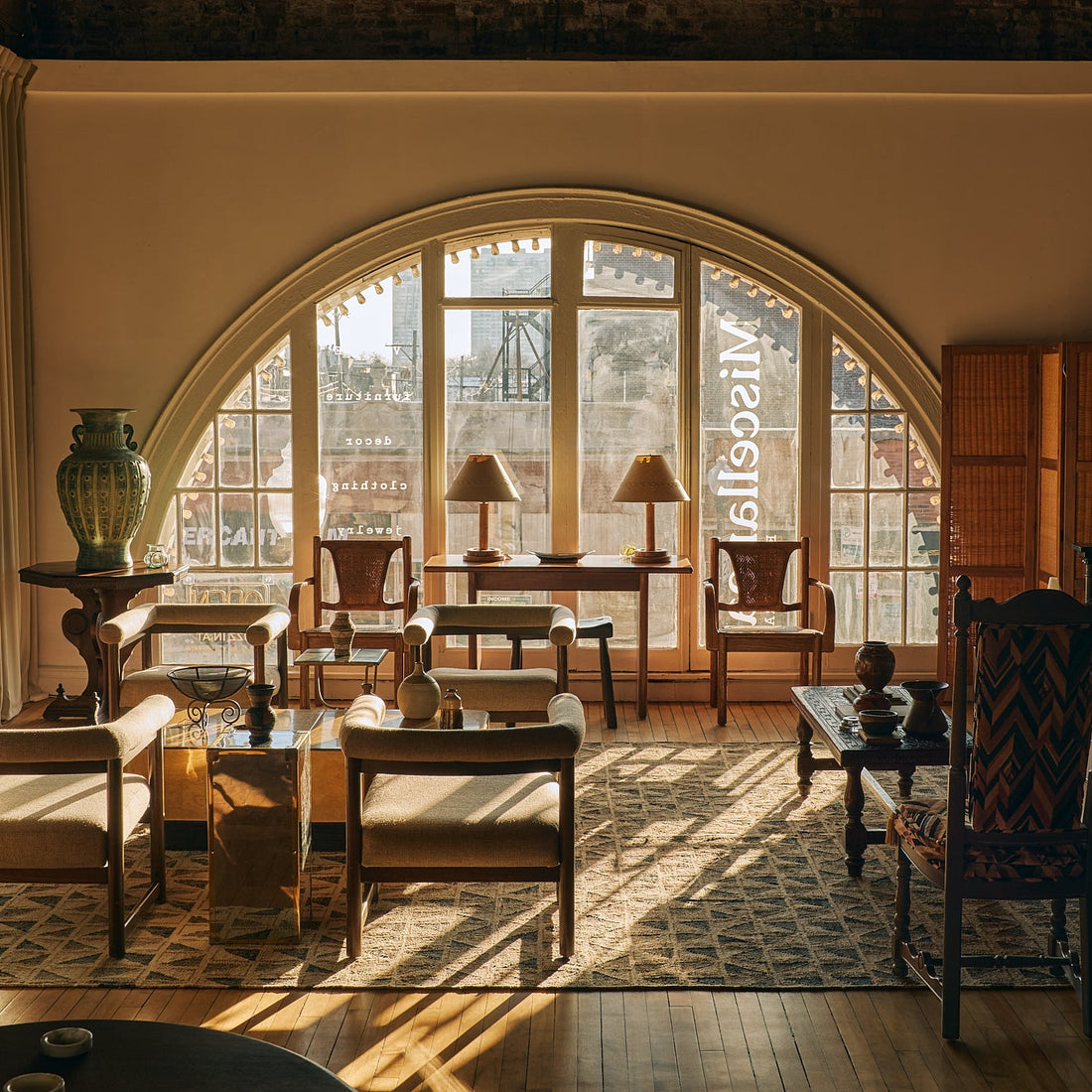 A luxury interior design scene showcasing a beautifully styled living room with a large arched window, warm sunlight streaming through, and a collection of elegant mid-century modern furniture including armchairs, a sofa, wooden tables, and a patterned rug.