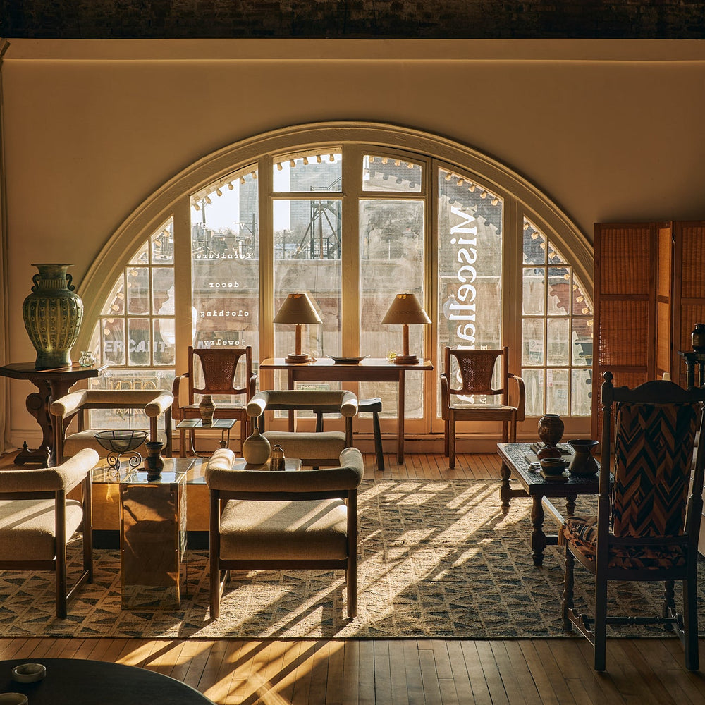 A luxury interior design scene showcasing a beautifully styled living room with a large arched window, warm sunlight streaming through, and a collection of elegant mid-century modern furniture including armchairs, a sofa, wooden tables, and a patterned rug.