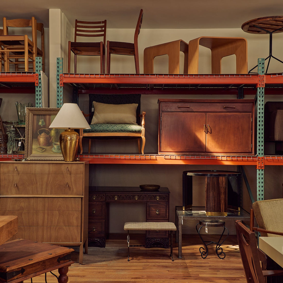 A vintage-style wooden furniture showroom or warehouse interior with multiple pieces of furniture displayed on metal shelving. Visible items include wooden chairs, a cabinet, a desk, a stool, and tables, arranged in a warm, rustic retail environment.