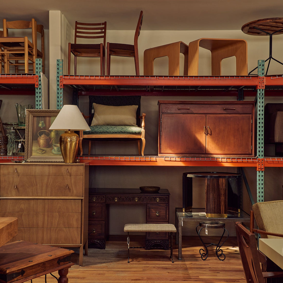 A vintage-style wooden furniture showroom or warehouse interior with multiple pieces of furniture displayed on metal shelving. Visible items include wooden chairs, a cabinet, a desk, a stool, and tables, arranged in a warm, rustic retail environment.