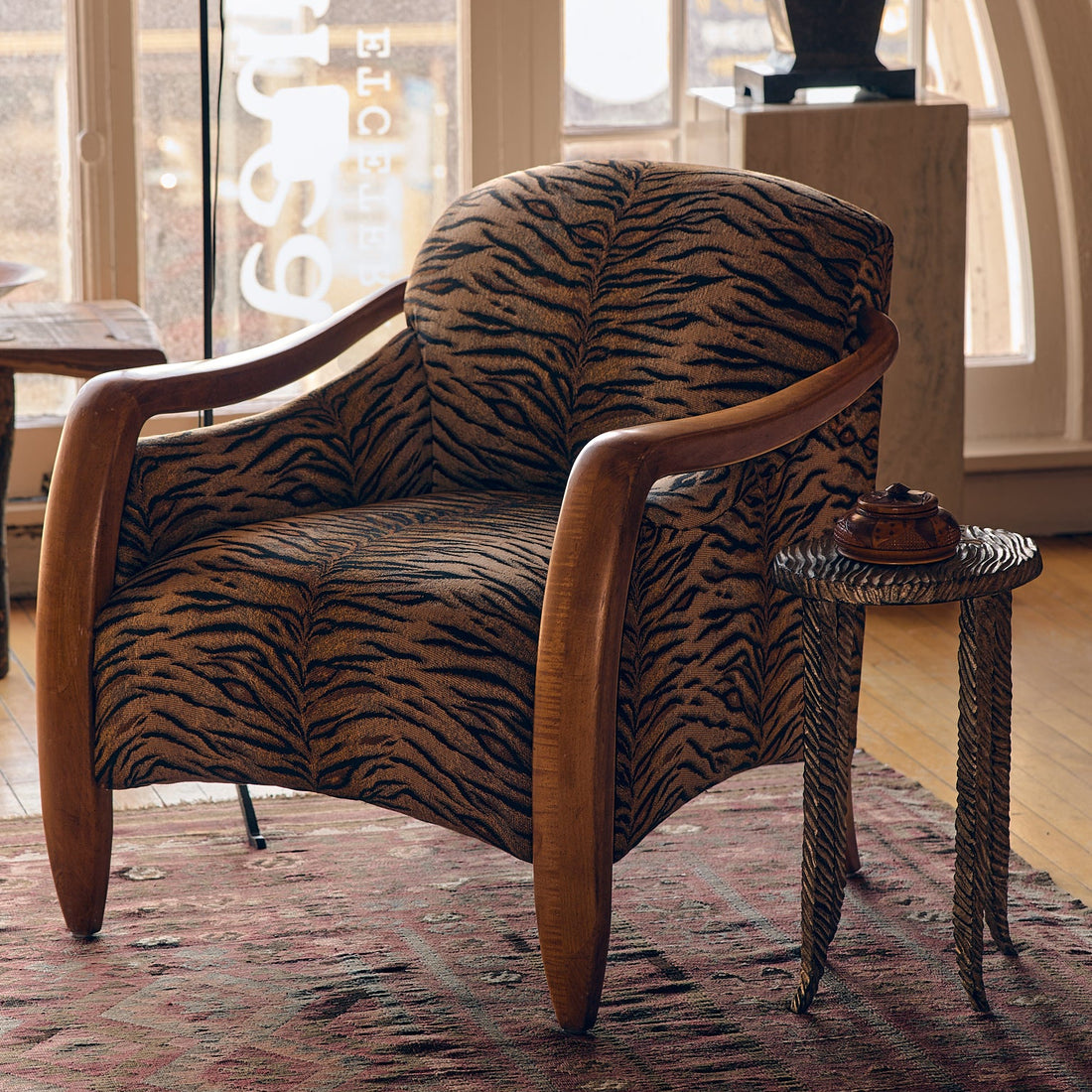 A modern armchair with a brown and black tiger-stripe patterned fabric and curved wooden arms, styled in a warm, sunlit room next to a small side table.