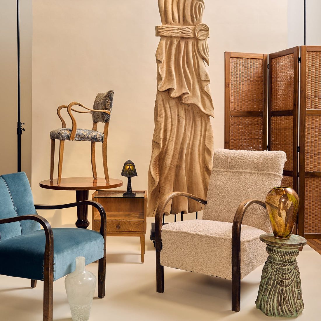 A styled interior scene with multiple furniture pieces and decor: a blue upholstered armchair, a cream bouclé armchair, a wooden chair, a tall carved wooden statue, a folding screen, side tables, and a glass vase, arranged against a beige backdrop.