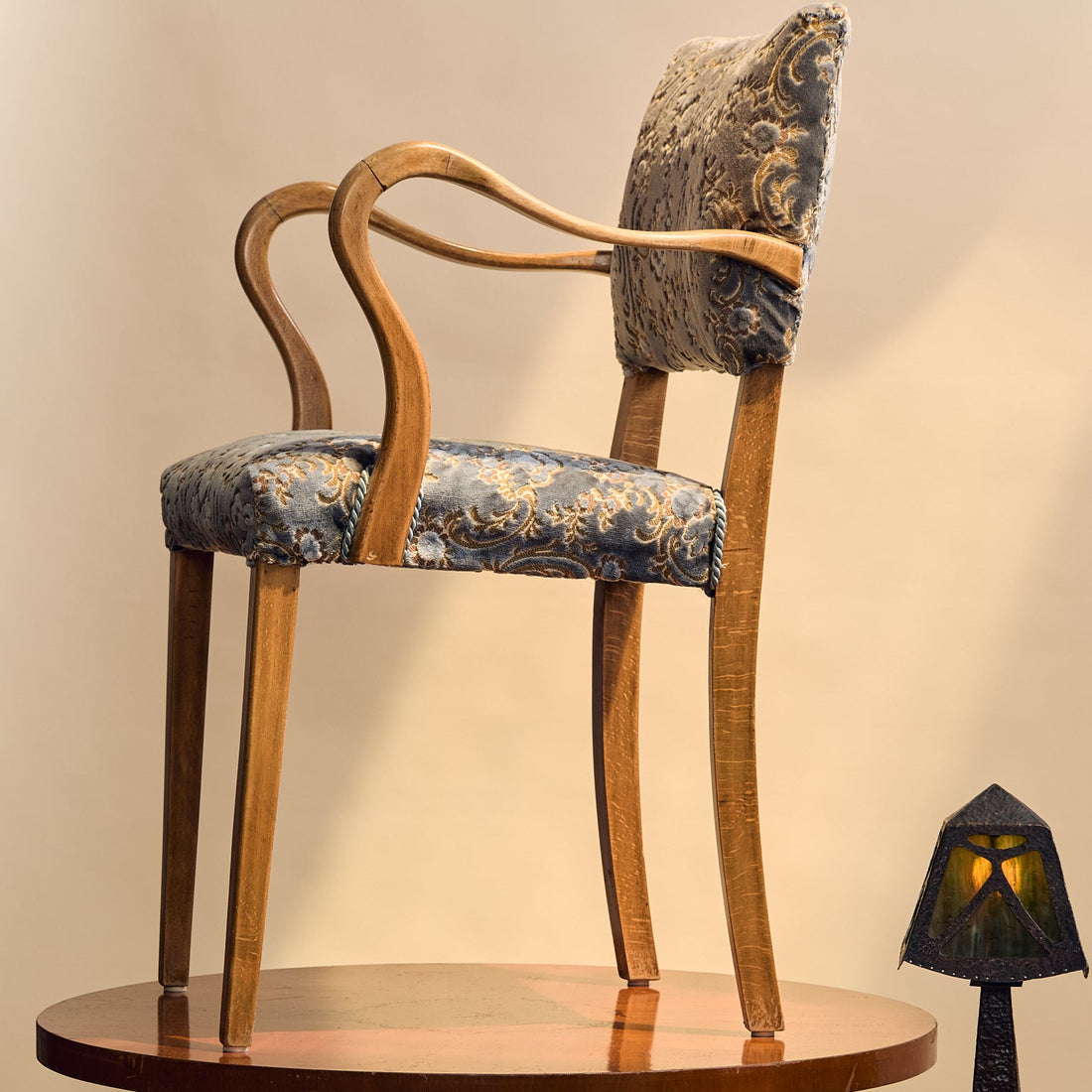 An elegant Art Deco style armchair with curved wooden arms and legs, upholstered in a blue and gold damask patterned fabric, displayed on a round wooden pedestal table.